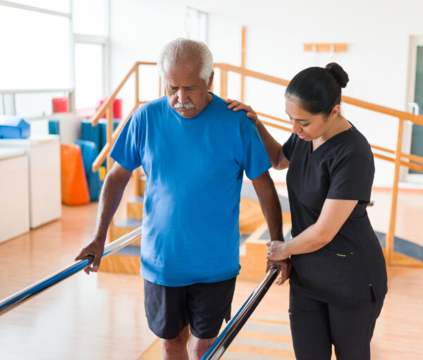 A female nurse helping a senior man to walk in the parallel bars.